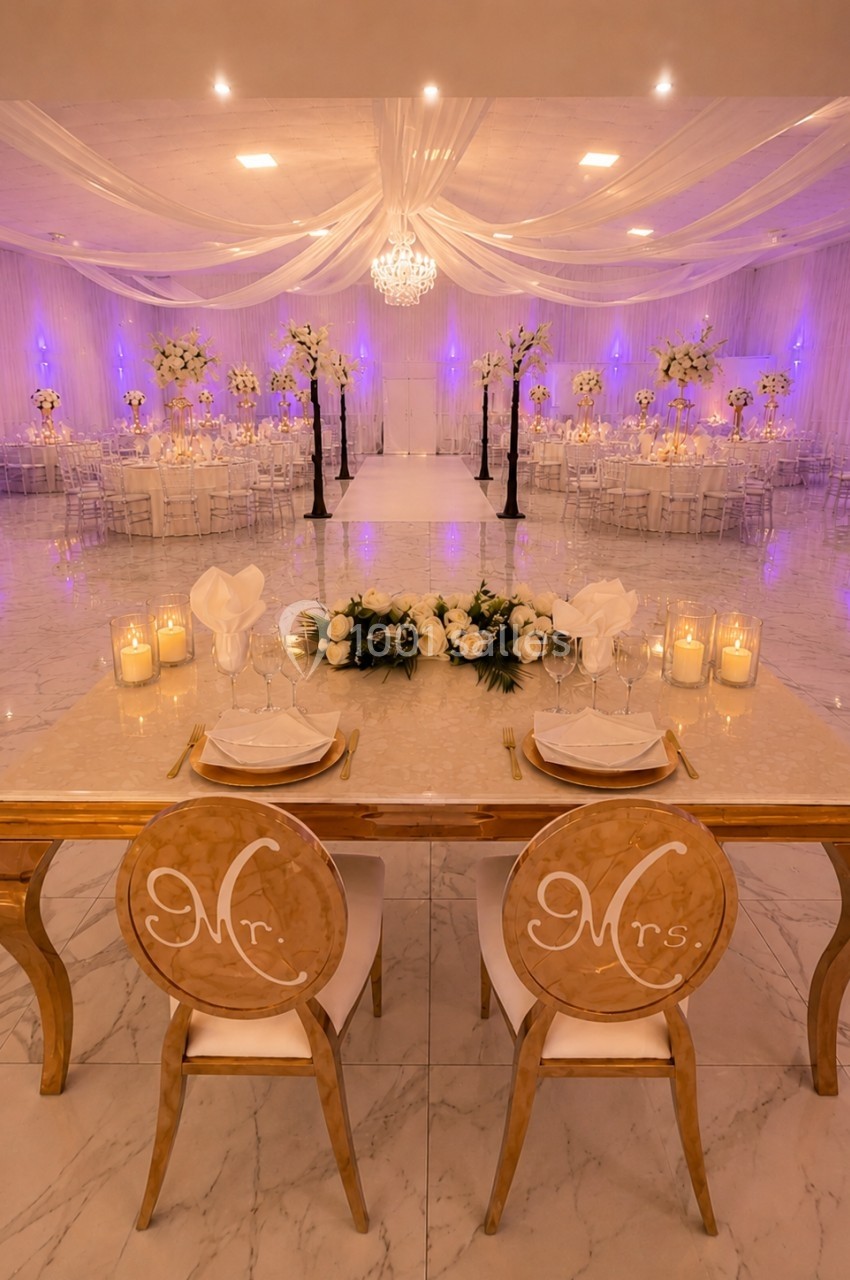 Salle de réception élégante avec table des mariés ornée de fleurs blanches, chandelles et décorations lumineuses.
