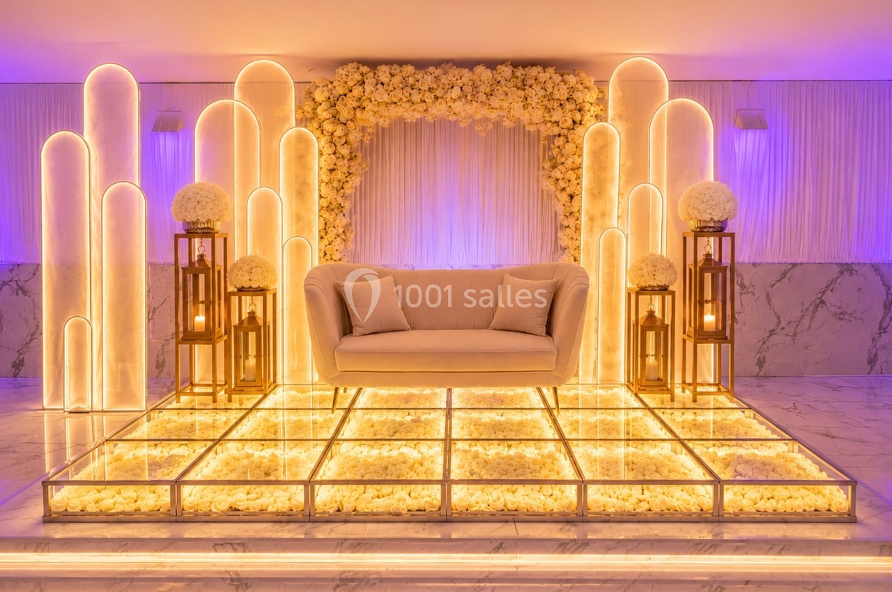 Décor lumineux avec canapé beige, arche florale blanche et lanternes dorées sur une scène en verre illuminée.