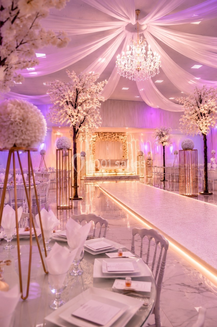 Salle de réception élégante décorée de fleurs blanches, éclairage rose et lustre central sous un plafond drapé.