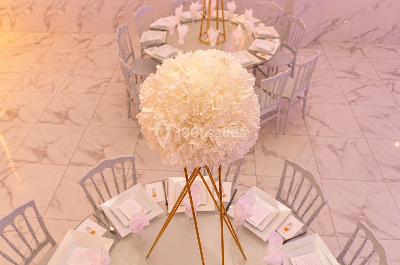 Centre de table floral blanc sur trépied doré, entouré de tables rondes dressées avec nappes blanches et chaises grises.