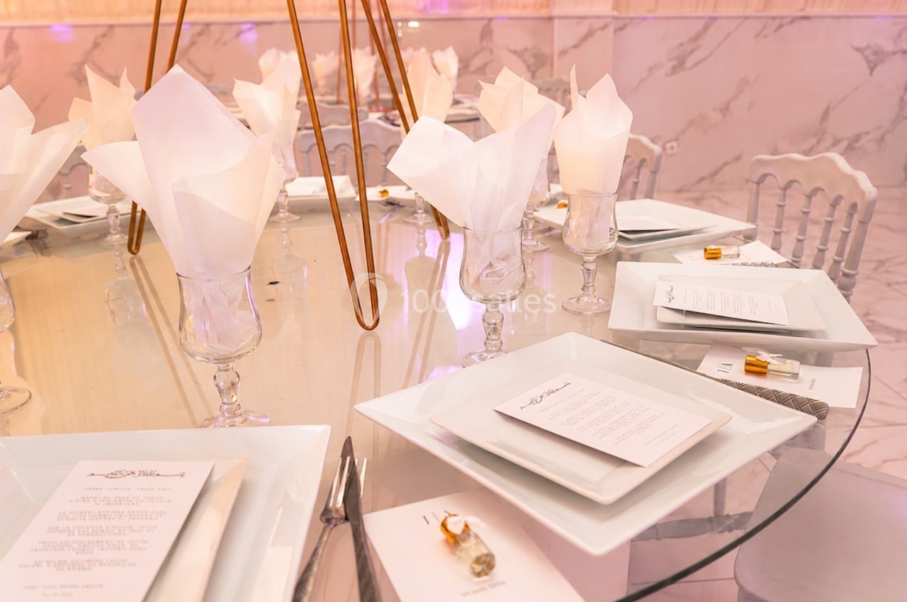 Table dressée avec des assiettes blanches, menus, serviettes pliées et décoration élégante sur une surface en verre.