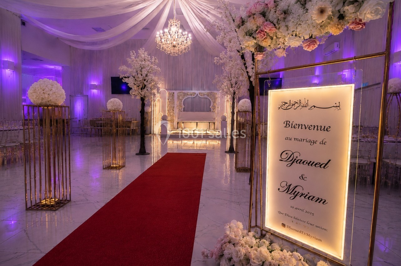 Salle de réception décorée pour un mariage avec tapis rouge, fleurs blanches et panneau de bienvenue illuminé.