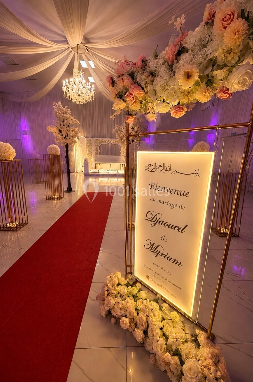 Entrée décorée d'une salle de mariage avec tapis rouge, fleurs, panneau lumineux et éclairage tamisé.
