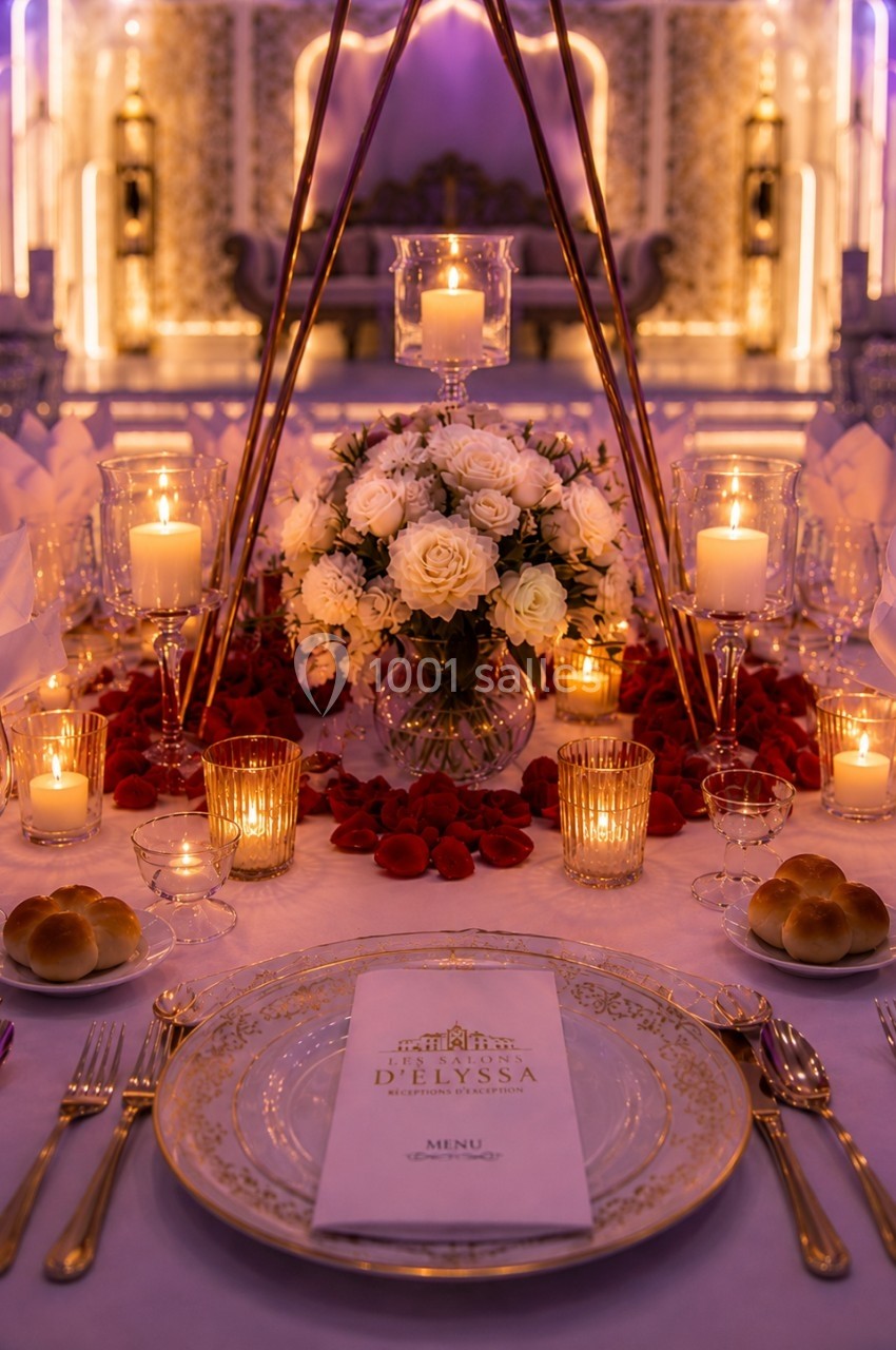 Table élégamment dressée avec des bougies, un bouquet de fleurs blanches et des pétales rouges, dans une ambiance festive.