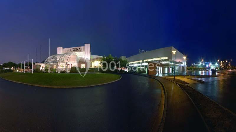 Vue nocturne d'un bâtiment moderne éclairé, entouré d'espaces verts et de routes humides reflétant les lumières.