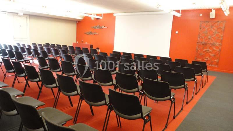 Salle de conférence vide avec des rangées de chaises noires, un écran de projection blanc et un mur rouge en arrière-plan.