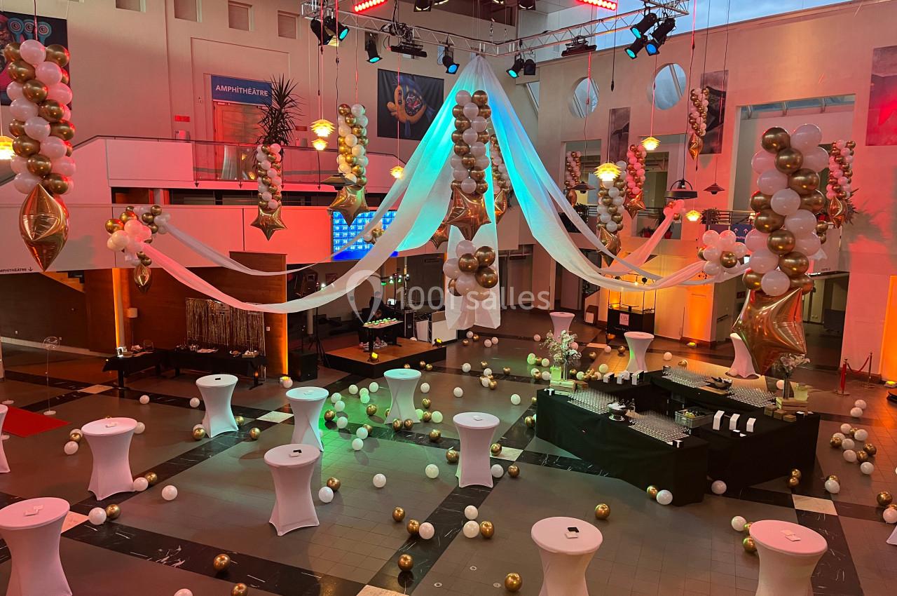 Salle décorée pour un événement avec ballons dorés et blancs, tables hautes et éclairages colorés.