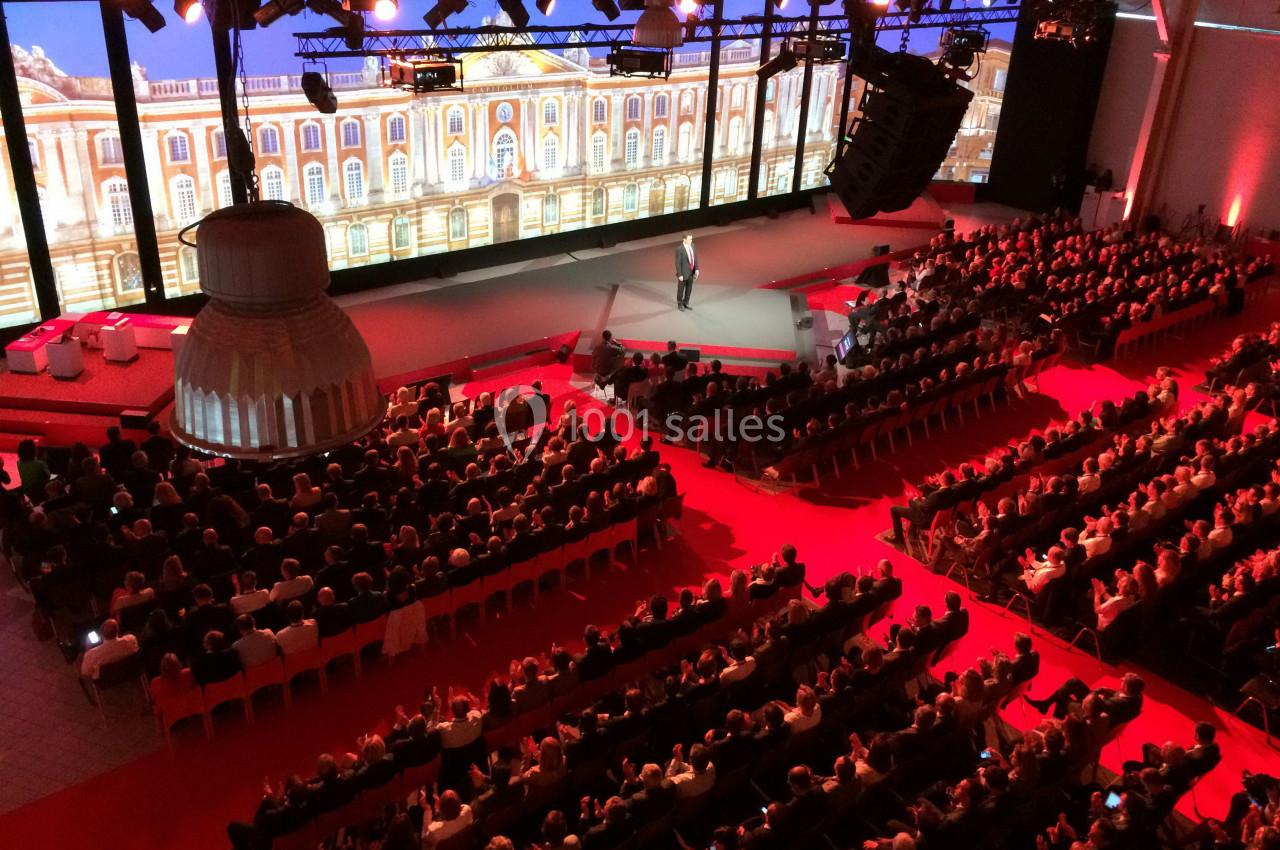 Salle de conférence avec un public nombreux, scène éclairée et écran géant affichant une façade architecturale.