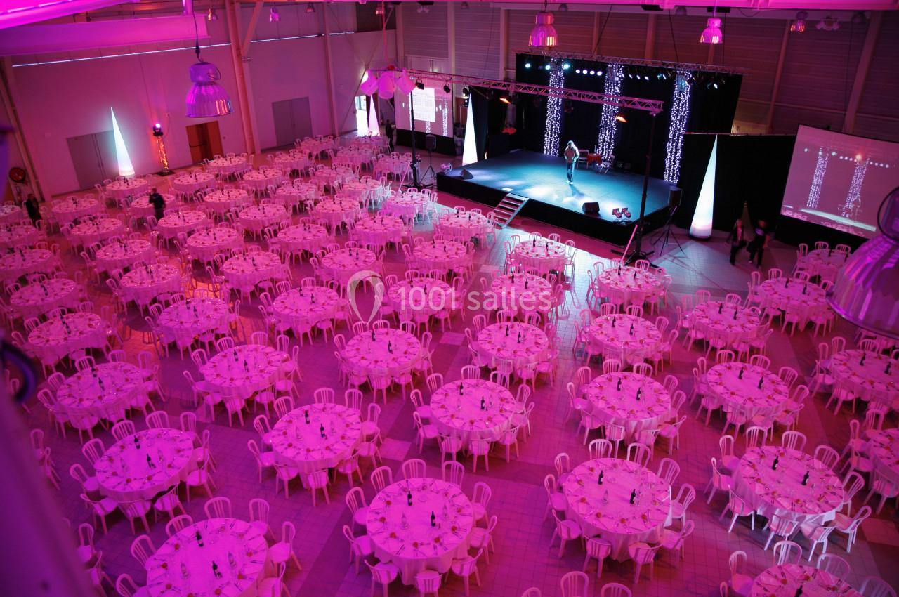 Salle de réception illuminée en rose avec de nombreuses tables rondes dressées et une scène au fond.