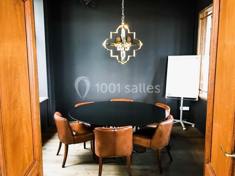 Salle de réunion avec table ronde noire, chaises en cuir marron, mur sombre, tableau blanc et suspension lumineuse.