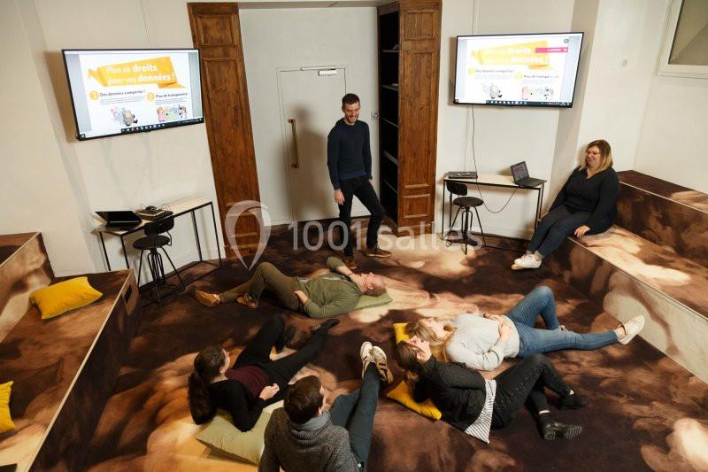 Un groupe de personnes échange dans une salle équipée d'écrans, certaines assises au sol sur des coussins.