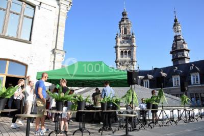 Personnes rassemblées sous une tente verte sur une terrasse, avec des plantes en pot et des clochers en arrière-plan.