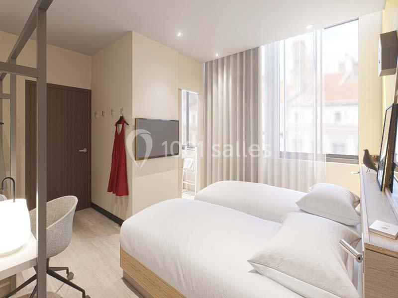 Chambre lumineuse avec deux lits simples, un bureau, une télévision murale et une robe rouge accrochée près de la porte.