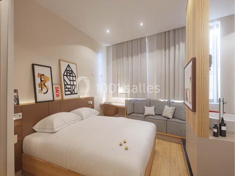 Chambre moderne avec lit double, canapé, décor minimaliste et lumière naturelle filtrée par des rideaux.