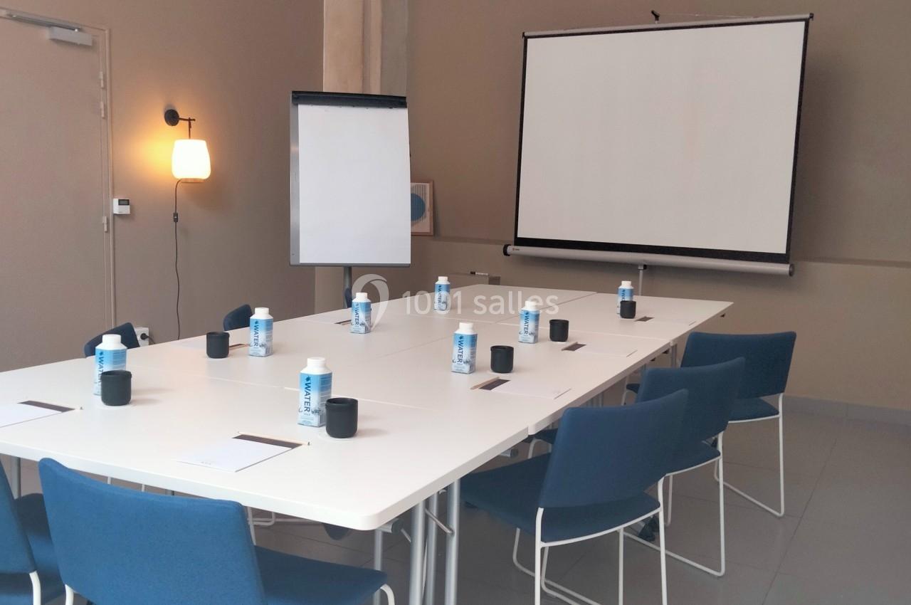 Salle de réunion avec table rectangulaire, chaises bleues, paperboard, vidéoprojecteur et bouteilles d'eau disposées.