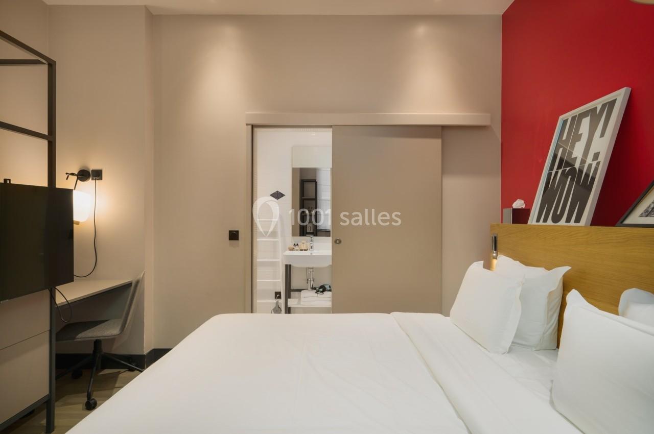 Chambre d'hôtel moderne avec lit double, bureau, télévision, et salle de bain visible à travers une porte coulissante.