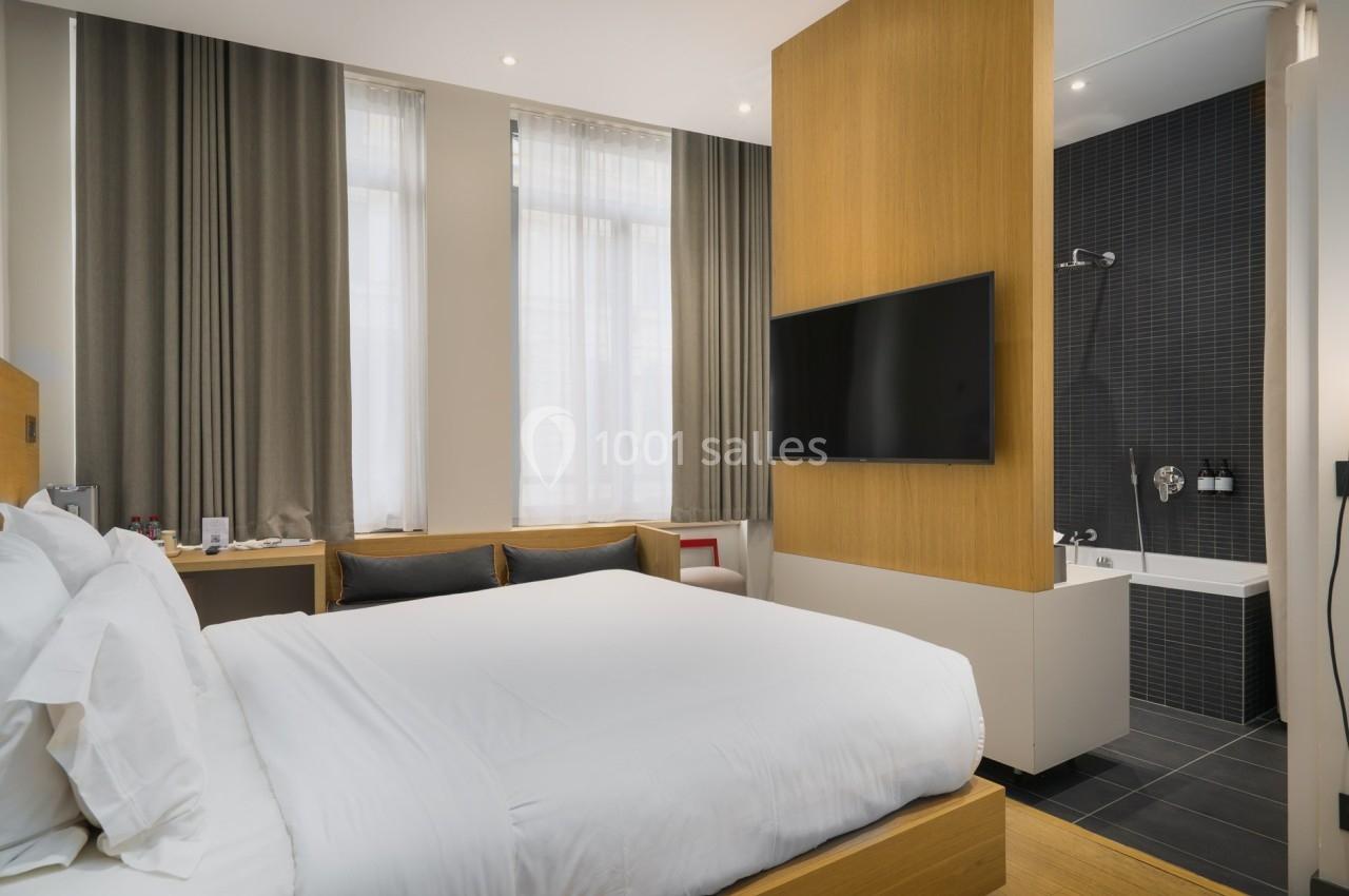 Chambre d'hôtel moderne avec lit double, télévision murale, grande fenêtre et salle de bain attenante avec baignoire.