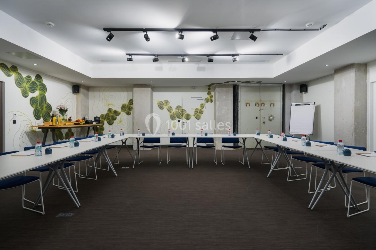 Salle de réunion lumineuse avec tables disposées en U, chaises bleues, tableau de conférence et buffet en arrière-plan.