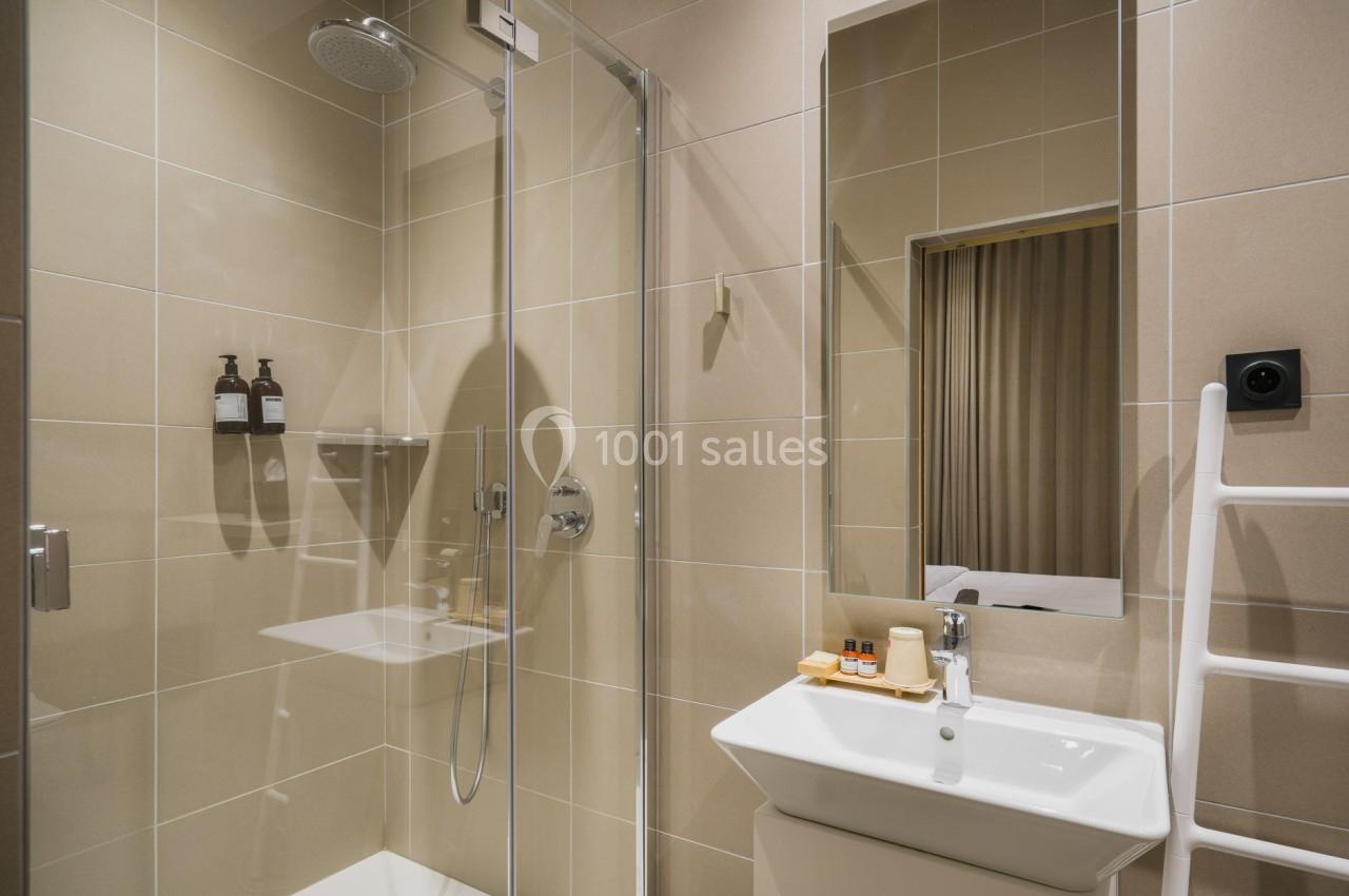 Salle de bain moderne avec douche vitrée, lavabo blanc, miroir, et accessoires de toilette sur un mur carrelé beige.