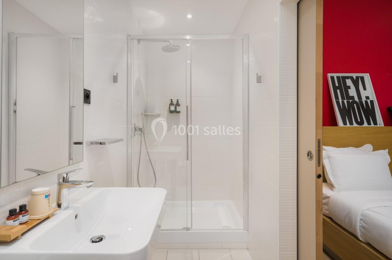 Salle de bain moderne avec douche vitrée, lavabo blanc et accessoires, partiellement ouverte sur une chambre.