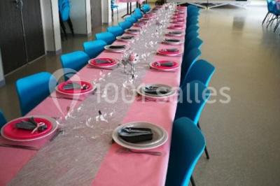 Salle lumineuse avec deux rangées de tables et chaises bleues, grandes fenêtres et portes vitrées sur les côtés.
