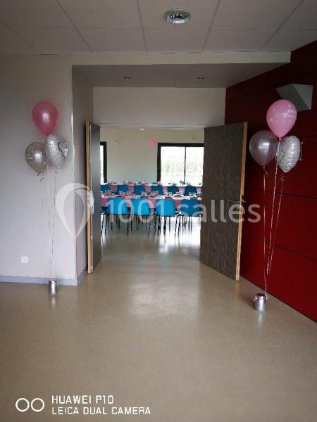 Salle décorée pour un événement, avec des ballons à l'entrée et des tables dressées visibles en arrière-plan.