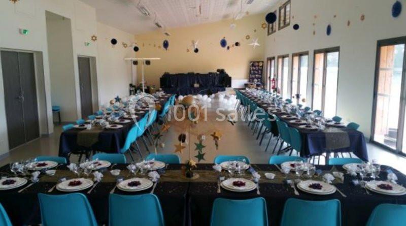 Salle décorée pour un événement, avec de longues tables dressées, chaises bleues et guirlandes suspendues.