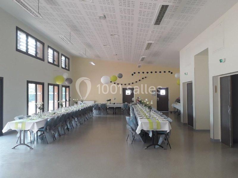 Salle décorée avec des tables alignées et dressées pour un événement, ornée de guirlandes et de suspensions colorées.