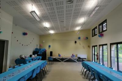 Salle lumineuse avec deux rangées de tables et chaises bleues, grandes fenêtres et portes vitrées sur les côtés.
