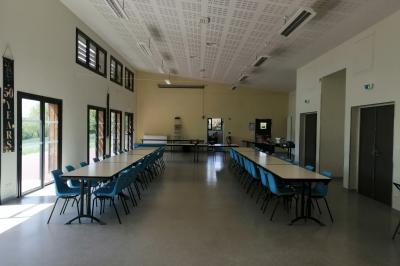 Salle lumineuse avec deux rangées de tables et chaises bleues, grandes fenêtres et portes vitrées sur les côtés.