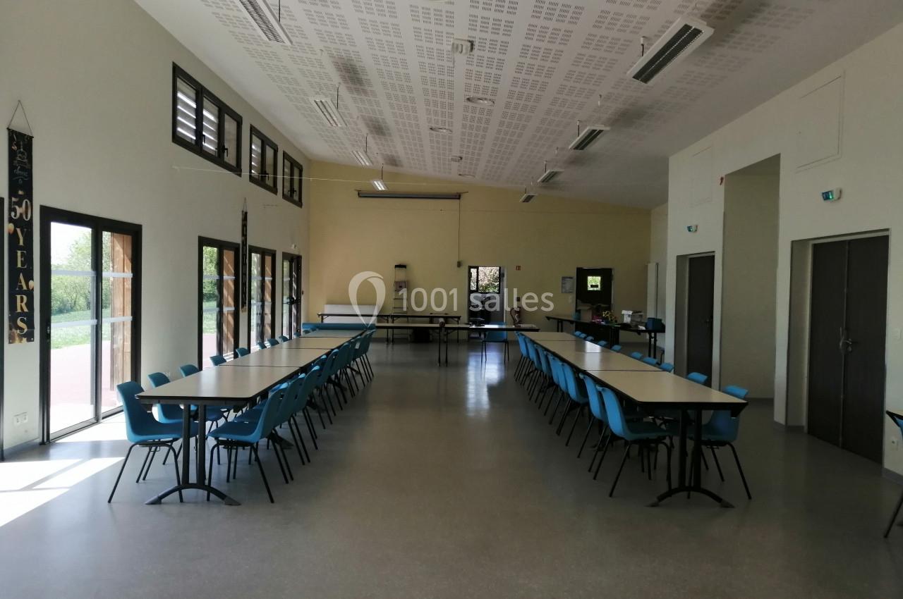 Salle lumineuse avec deux rangées de tables et chaises bleues, grandes fenêtres et portes vitrées sur les côtés.