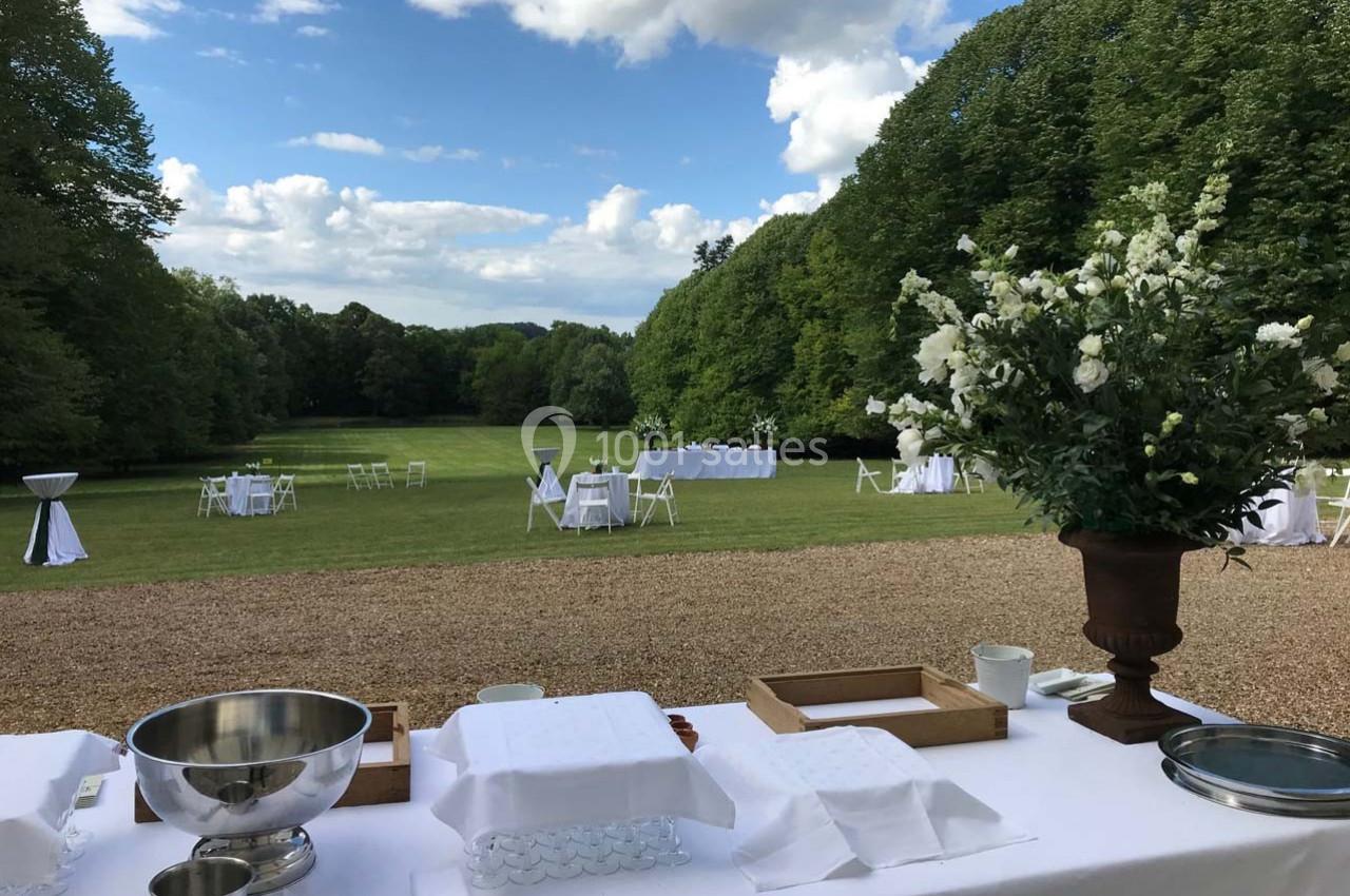 Table dressée en extérieur avec vue sur un grand jardin verdoyant, des tables blanches dispersées au loin.