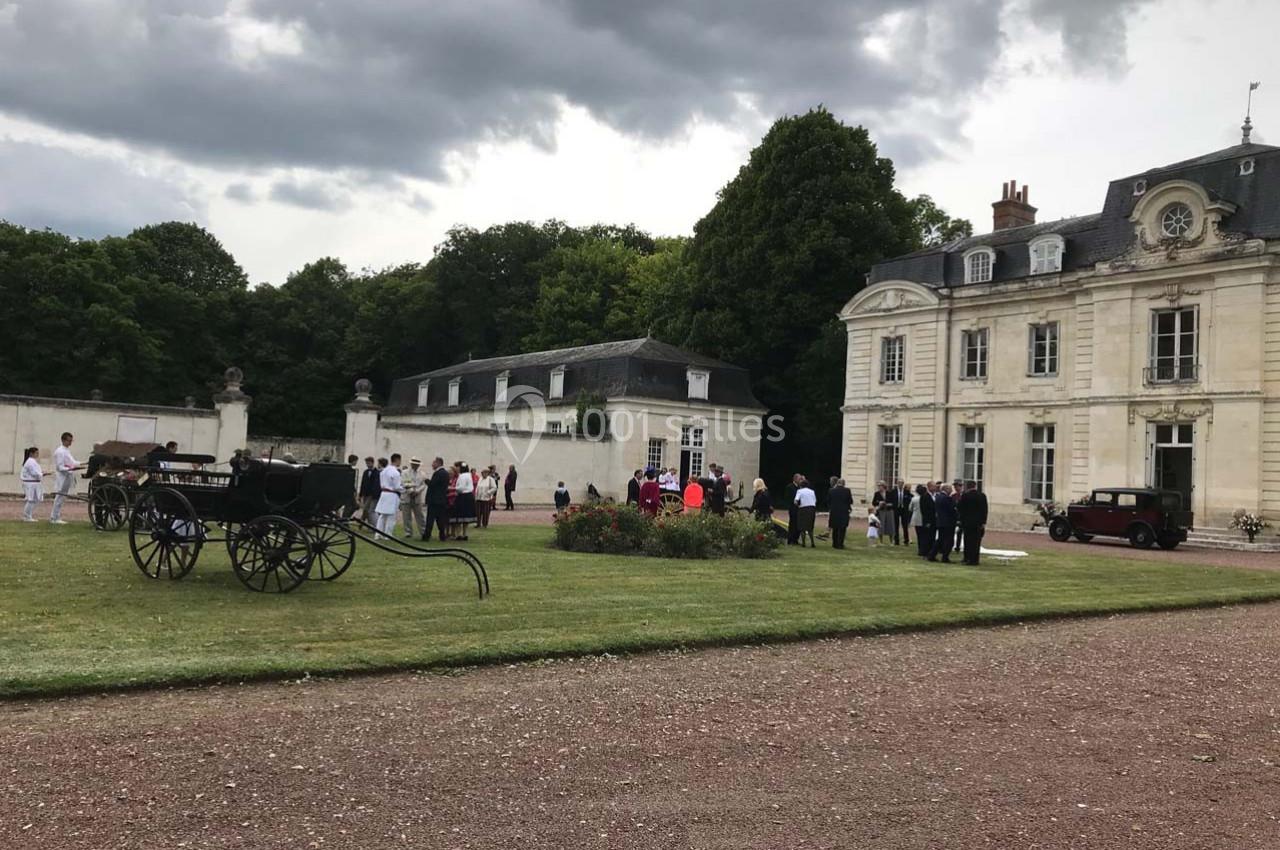 Groupe de personnes rassemblées dans la cour d'un château entouré de verdure, avec des calèches en premier plan.