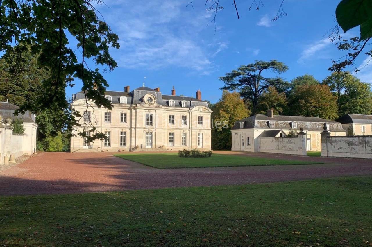 Façade d'un château entouré de dépendances, avec une allée en gravier et un parc arboré sous un ciel bleu.