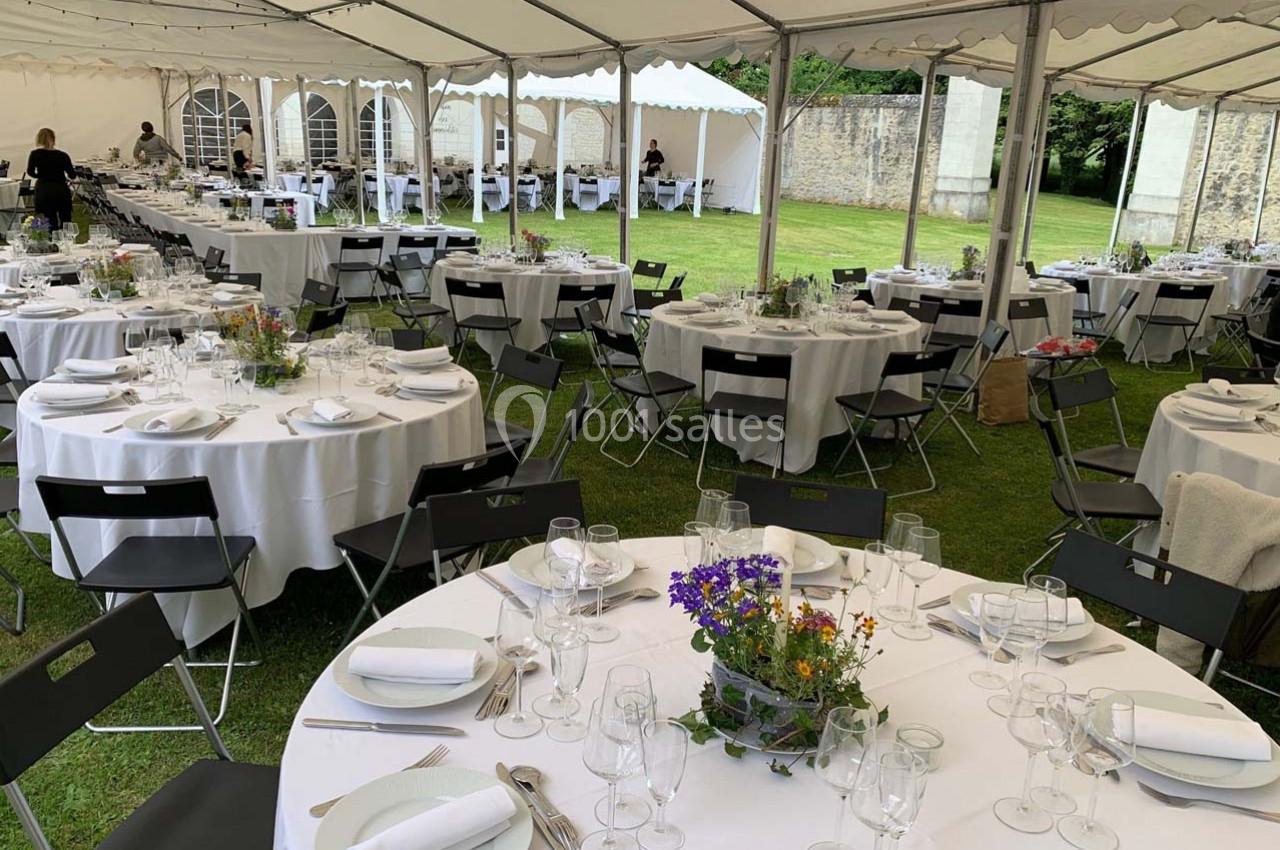 Tables rondes dressées avec nappes blanches, couverts et décorations florales sous une grande tente en extérieur.