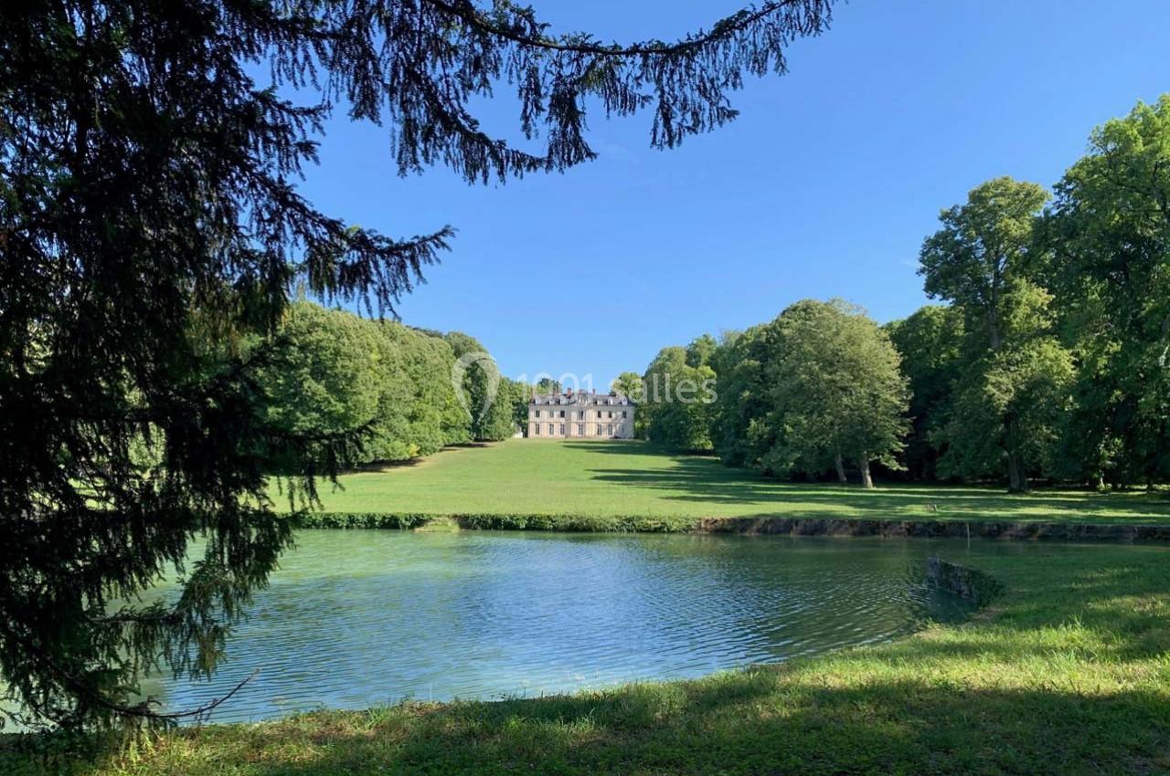Vue d'un château au loin, entouré de vastes pelouses et d'arbres, avec un étang au premier plan.