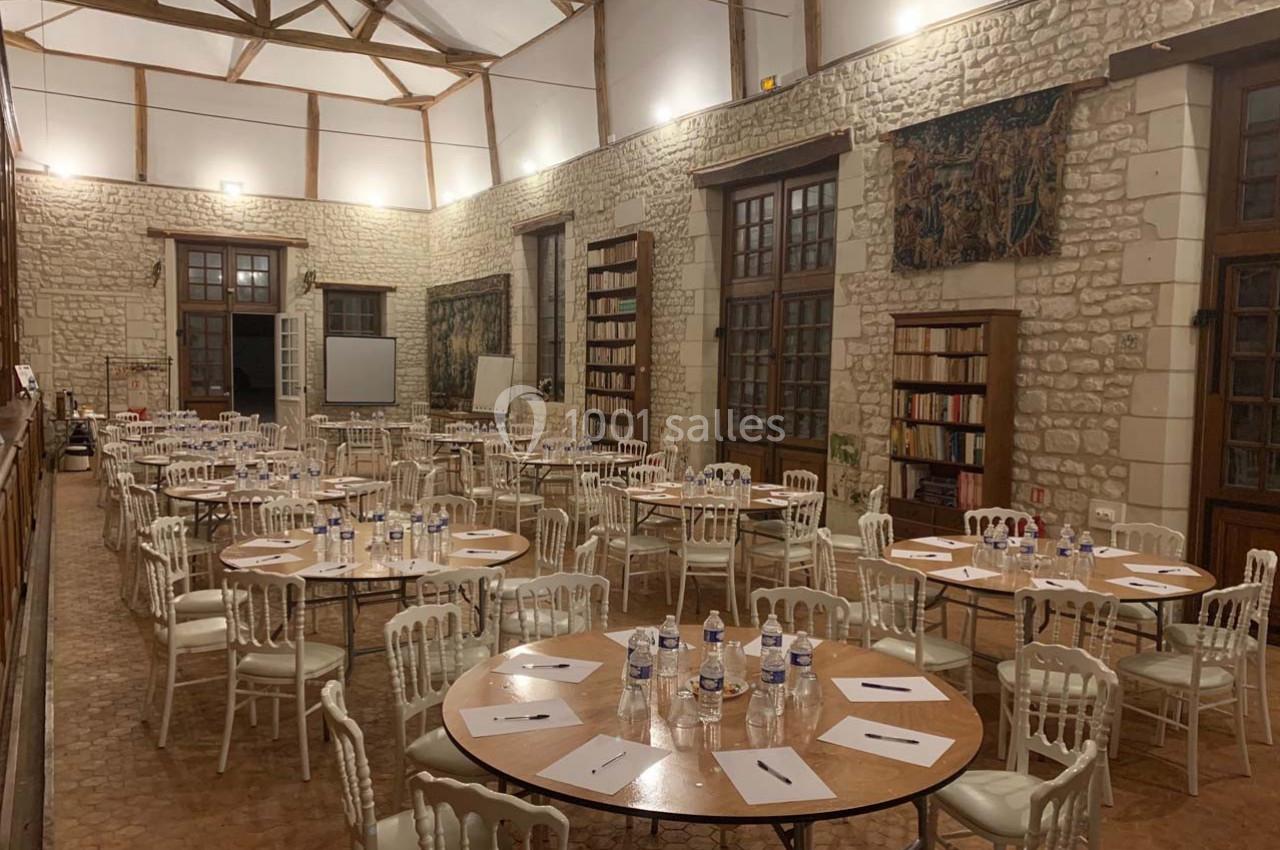 Salle en pierre avec des tables rondes dressées, chaises blanches, bouteilles d'eau et documents, ambiance de réunion.