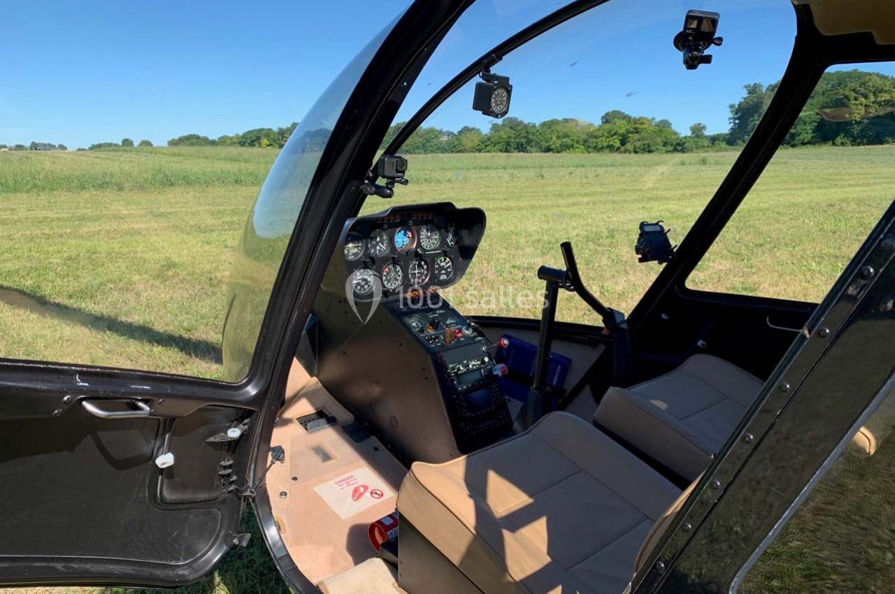Vue intérieure d'un hélicoptère avec tableau de bord, sièges beiges et paysage de prairie en arrière-plan.
