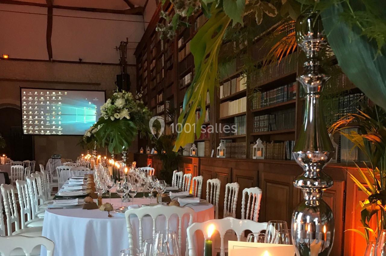 Salle de réception décorée avec des tables rondes, des chandeliers, des fleurs et une bibliothèque en arrière-plan.