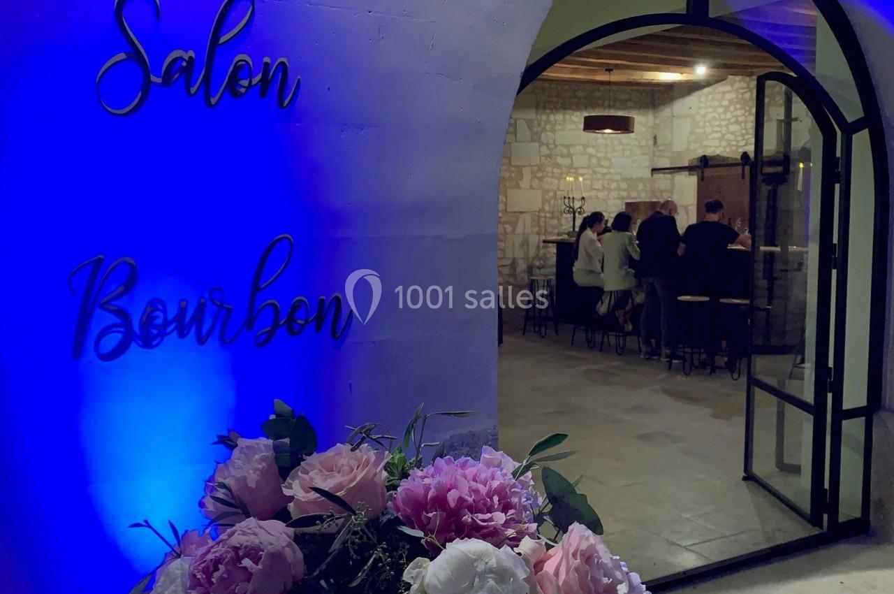 Entrée d'un salon éclairé en bleu, avec des fleurs au premier plan et des personnes discutant autour d'un bar à l'intérieur.