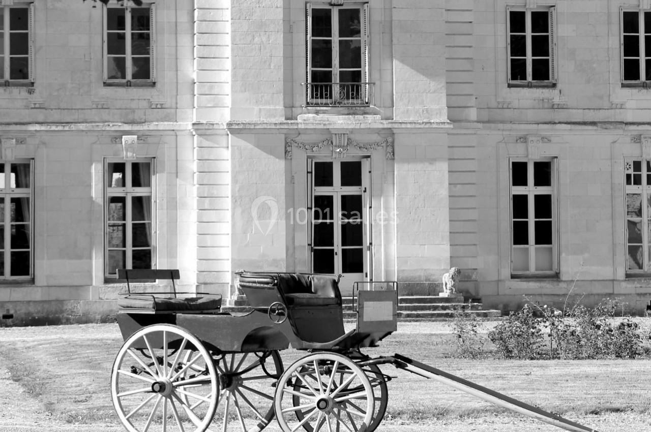 Calèche ancienne en bois avec roues blanches, stationnée devant une façade de bâtiment en pierre avec grandes fenêtres.