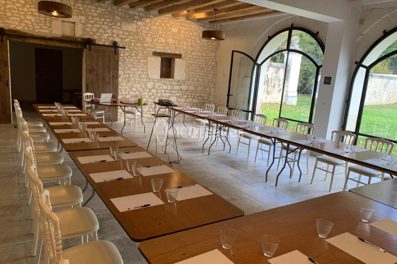 Salle lumineuse aménagée en configuration de réunion avec tables alignées, chaises blanches et grandes baies vitrées.