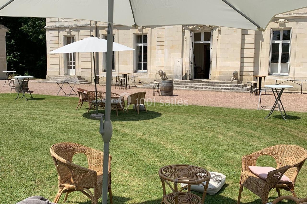 Mobilier de jardin en rotin disposé sous un parasol blanc sur une pelouse devant un bâtiment en pierre.