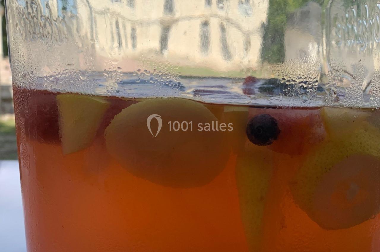 Bocal en verre contenant une boisson fruitée avec des morceaux de fruits, posé devant un bâtiment flou en arrière-plan.