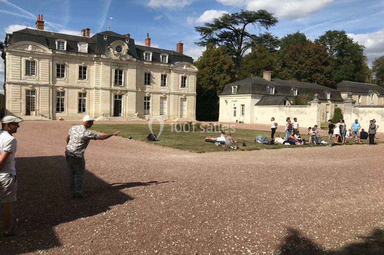 Un groupe de personnes joue à la pétanque devant un bâtiment ancien entouré de verdure.