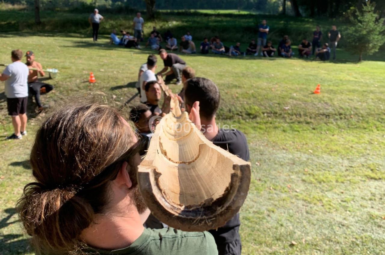 Deux personnes transportent une grande bûche en plein air, avec un groupe de spectateurs en arrière-plan.