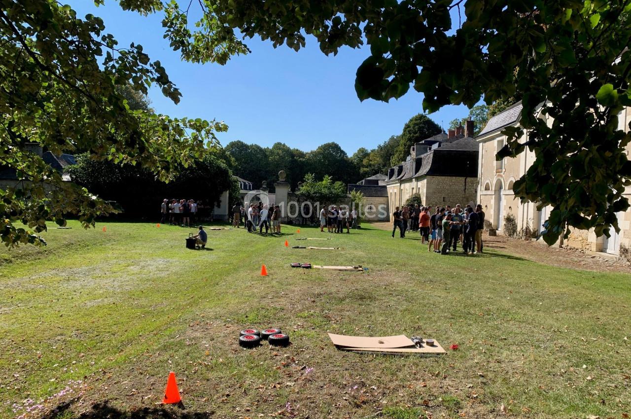 Groupe de personnes rassemblées dans une cour herbeuse près de bâtiments anciens, avec des activités au sol.