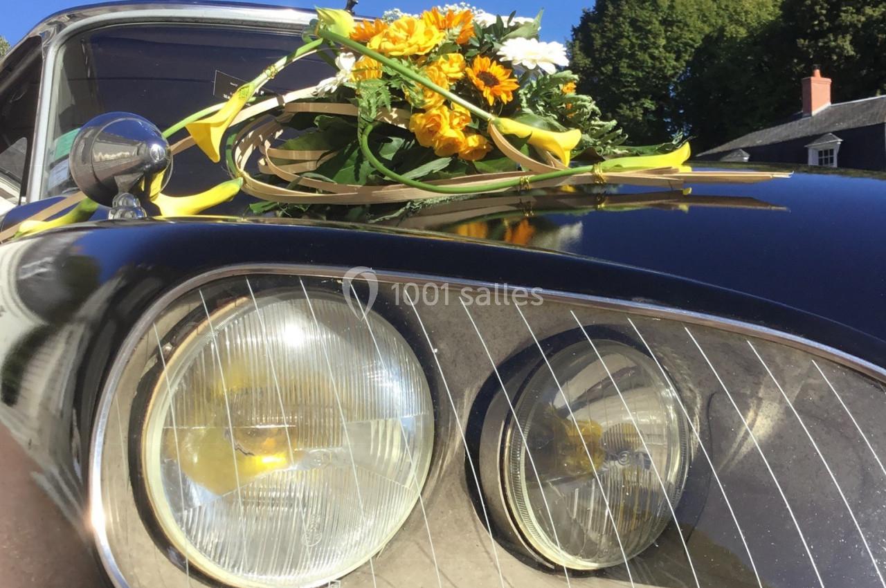 Bouquet de fleurs jaunes et blanches posé sur le capot d'une voiture ancienne noire, sous un ciel bleu.