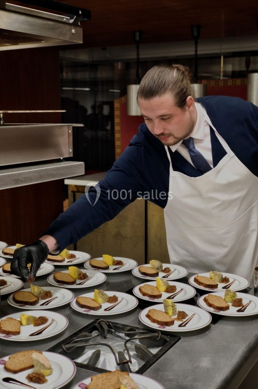 Un chef en tablier dispose des assiettes garnies de pain, fromage et accompagnements dans une cuisine professionnelle.