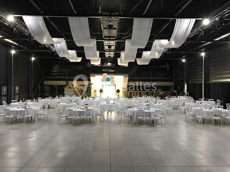 Salle de réception spacieuse avec des tables rondes dressées et des drapés blancs suspendus au plafond.